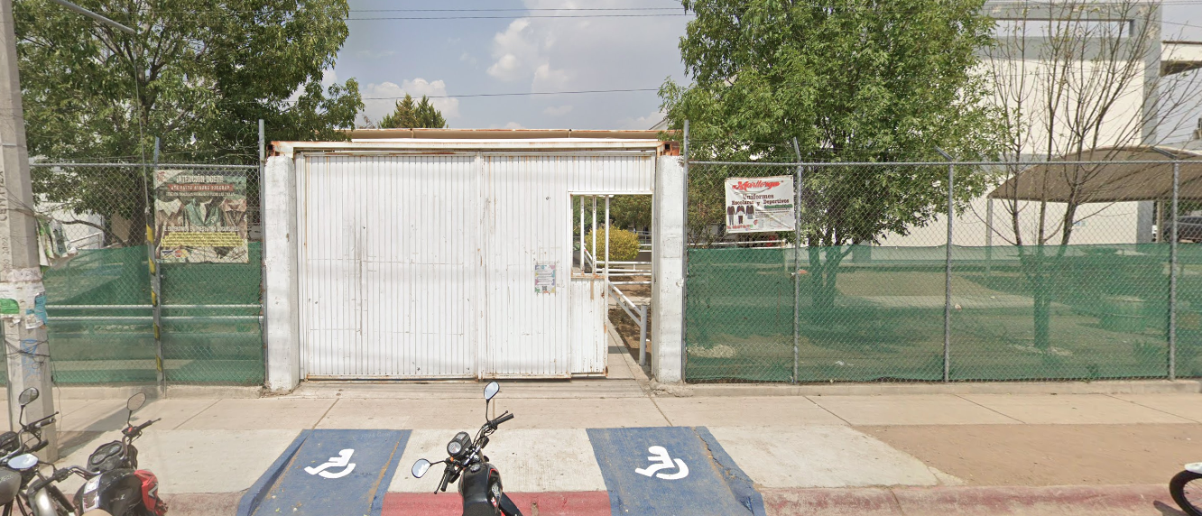 Fotografía donde se muestra la entrada del plantel conformada por un portón blanco con dos rampas de acceso en frente y el plantel está rodeado por una reja con malla color verde
